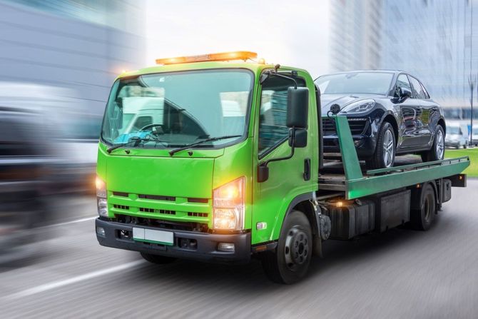Green flatbed tow truck carrying a black car on a blurred road