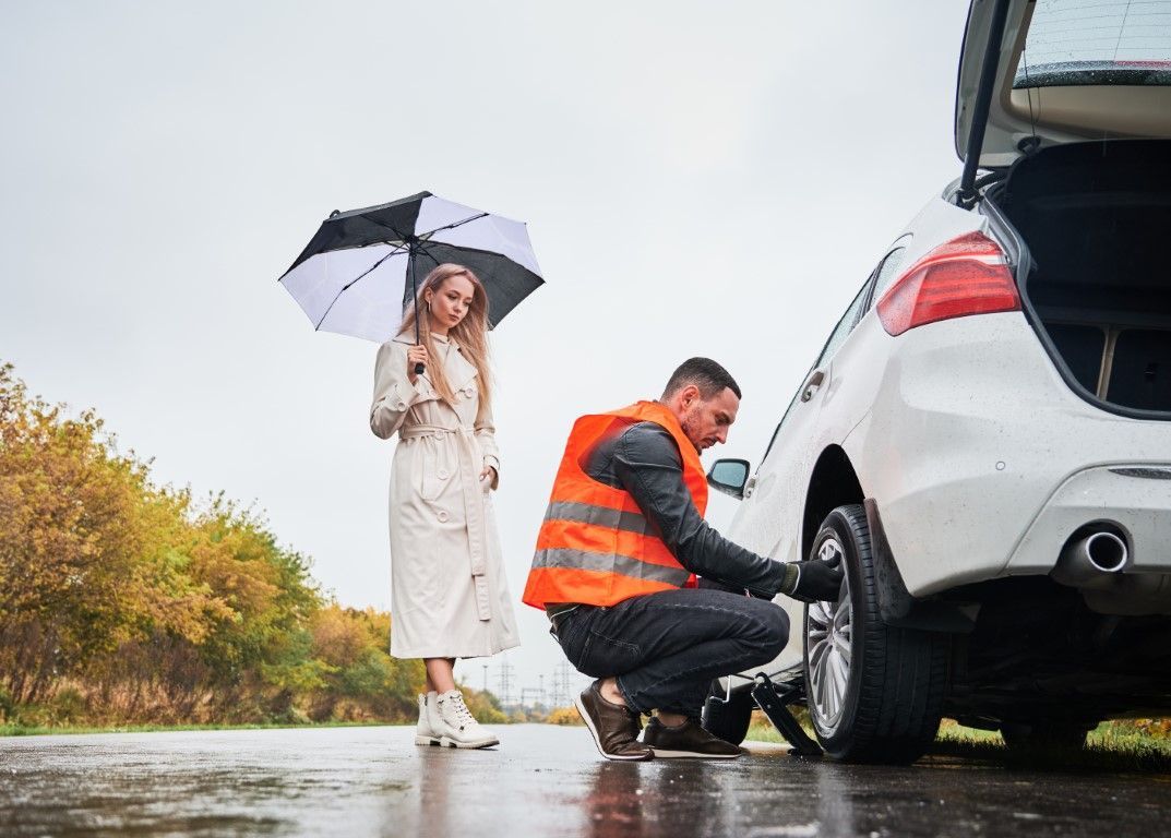 Mechanic changing a car tire in the rain while a person holds an umbrella nearby