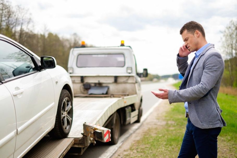 Tow truck loading a white car while a man talks on his phone beside the road